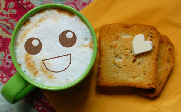 an overhead photo of a coffee cup and two slices of bread with a stick of heart-shaped butter. there is a happy face on top of the coffee.